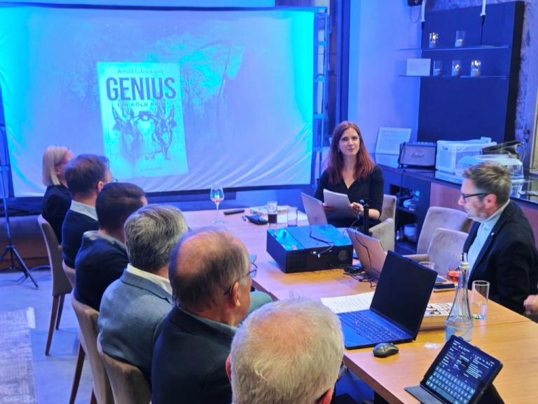 Frau am Tisch präsentiert ein Buch mit dem Titel "GENIUS" vor einer Gruppe von Zuhörern.