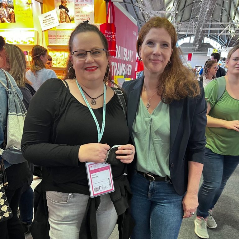 Zwei Frauen lächeln, stehen in einer belebten Umgebung mit Messeständen.