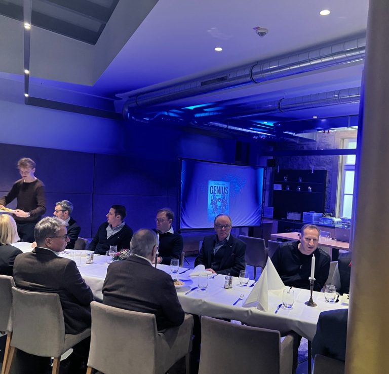 Person hält eine Rede vor einer Gruppe von Gästen in einem stilvollen Veranstaltungsraum.