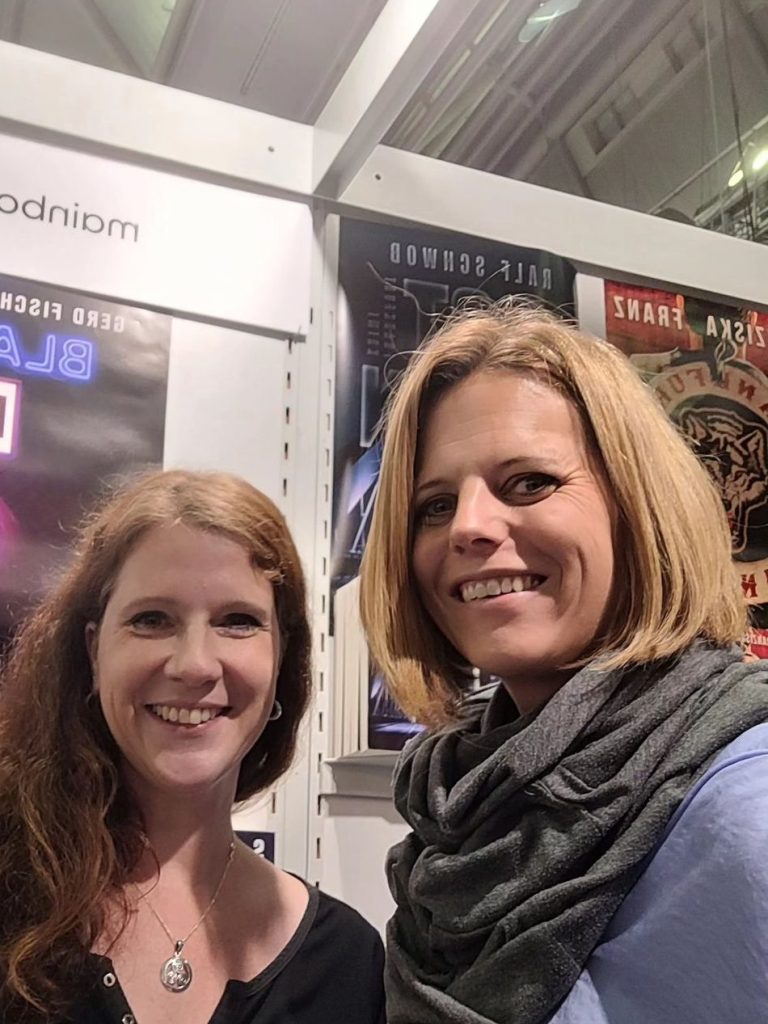 Zwei Frauen lächeln in die Kamera, stehen nebeneinander in einem Messeumfeld.