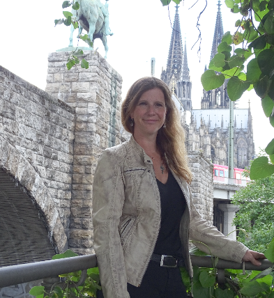 Porträt einer Frau mit langen, welligen Haaren, lächelnd vor einer historischen Steinmauer.