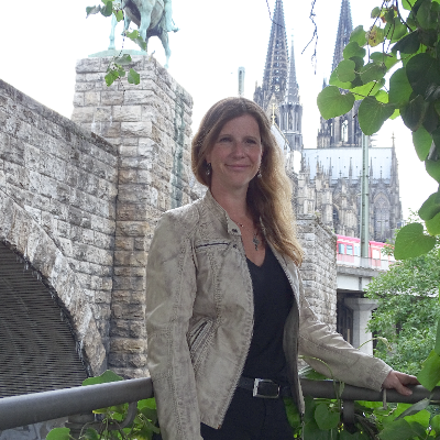 Porträt einer Frau mit langen, welligen Haaren, lächelnd vor einer historischen Steinmauer.