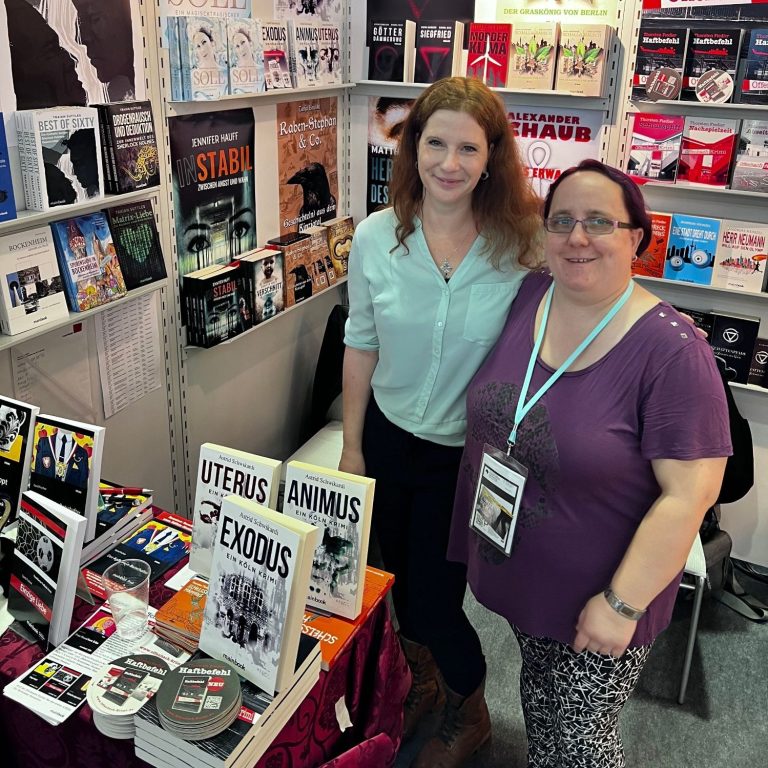 Zwei Frauen stehen an einem Messestand mit Büchern und Werbematerial.