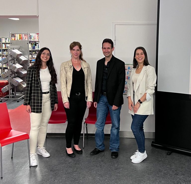 Vier Personen stehen in einem Raum mit Regalen, an einer Wand eine Leinwand.