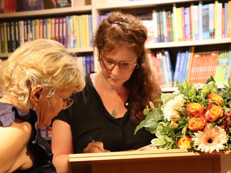 Eine Frau signiert ein Buch für eine Leserin in einer Buchhandlung. Blumenarrangement daneben.