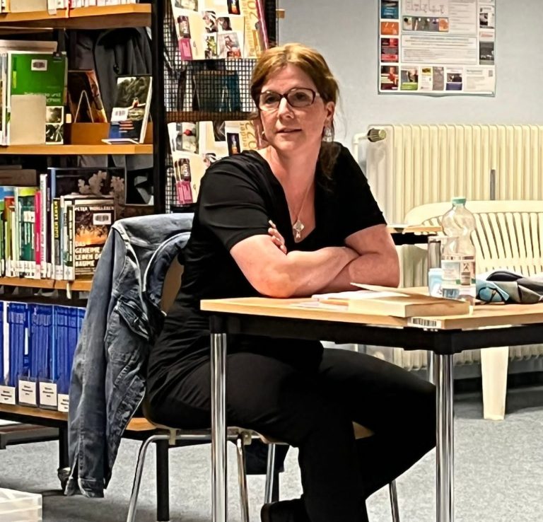 Frau mit Brille sitzt an einem Tisch in einer Bibliothek, lächelt und schaut auf.