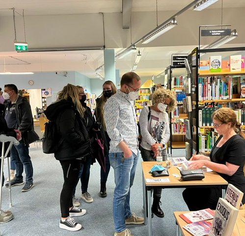Menschen stehen in einer Bibliothek, während eine Frau an einem Tisch beraten wird.