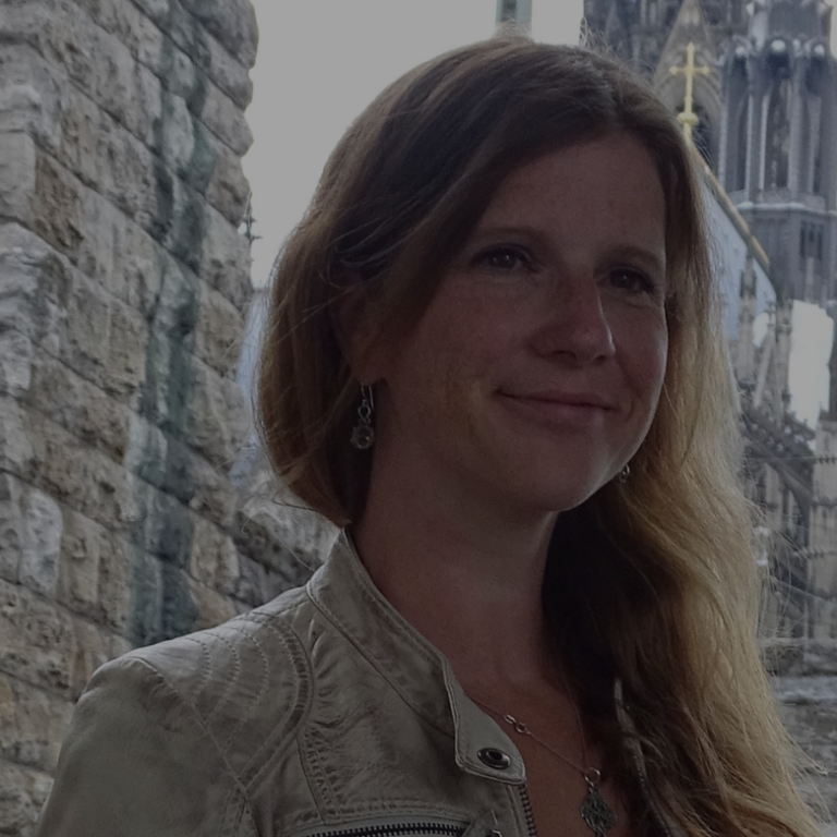 Porträt einer Frau mit langen, welligen Haaren, lächelnd vor einer historischen Steinmauer.