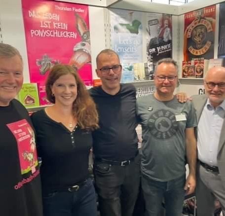 Fünf Personen stehen lächelnd vor bunten Plakaten und Buchcovern.