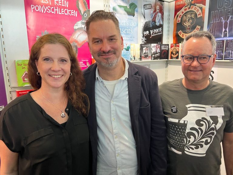 Drei Personen lächeln vor bunten Plakaten in einem hellen Raum.