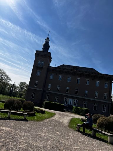 Historisches Gebäude mit Turm, umgeben von grünem Gelände und bänken.