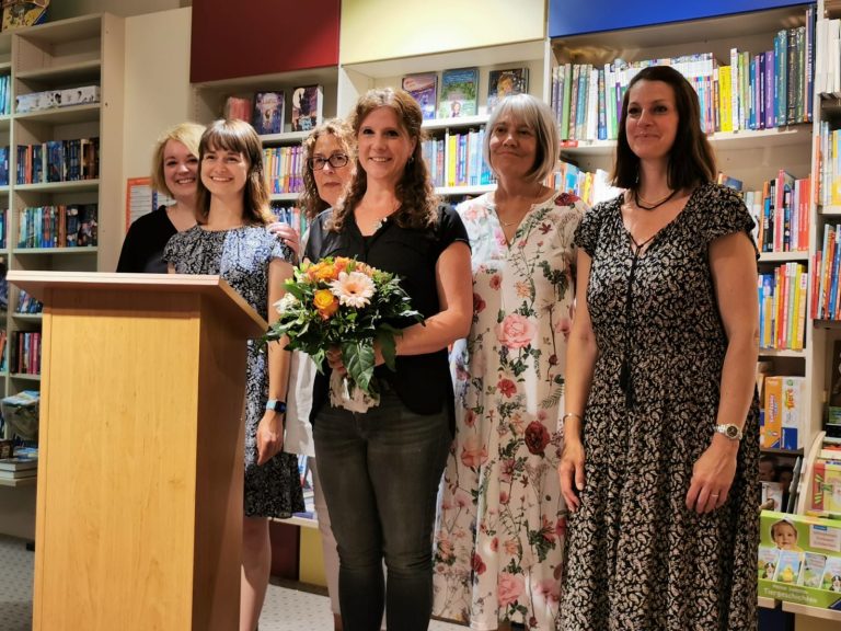 Sechs Frauen stehen lächelnd vor einem Bücherregal, eine hält einen Blumenstrauß.