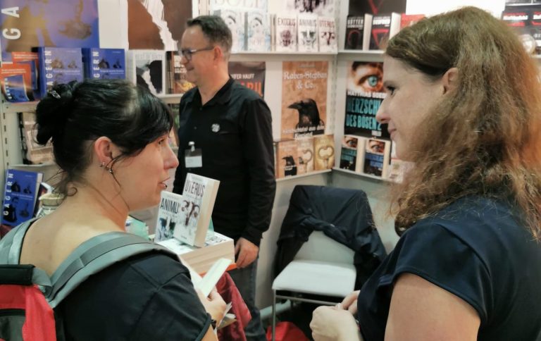 Zwei Frauen im Gespräch an einem Messestand, mit einem Mann im Hintergrund.