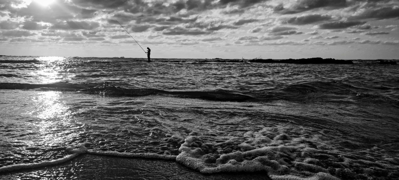 Schwarze und weiße Küstenlandschaft mit Wellen und einem Leuchtturm im Hintergrund.