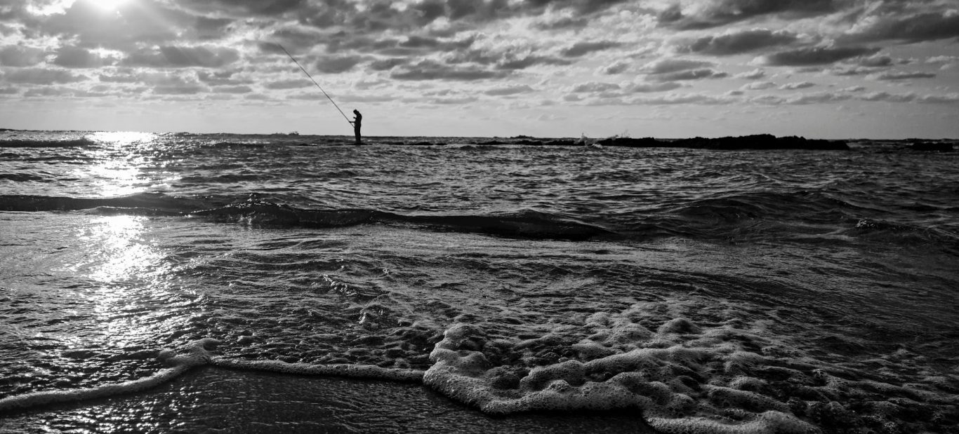 Schwarze und weiße Küstenlandschaft mit Wellen und einem Leuchtturm in der Ferne.
